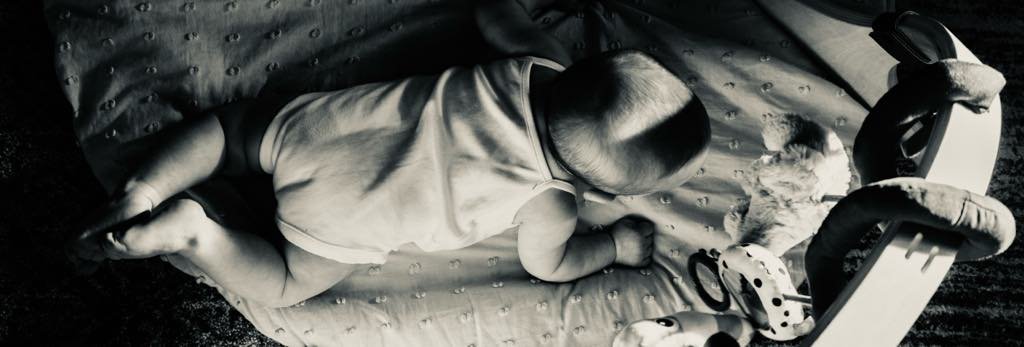Baby lying on tummy on a soft play mat during tummy time, surrounded by black-and-white toys and a plush animal demonstrating the benefits of tummy time.