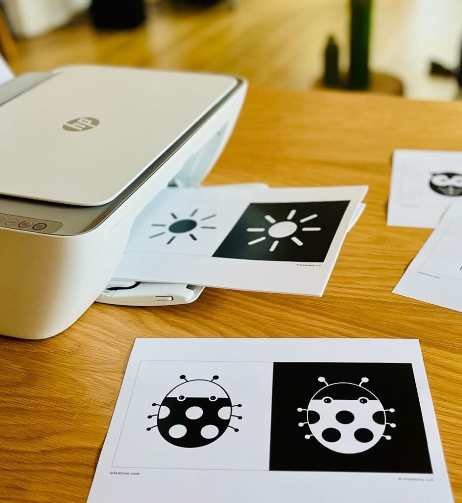 Printable black and white baby flashcards with high-contrast images emerging from a home printer on a wooden table.